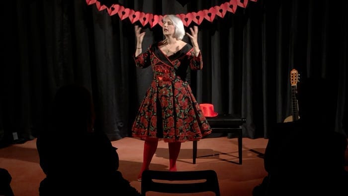 Christine Filip met rood/zwarte bloemetjes petticoat jurk op podium met de handen dramatisch omhoog. Het is een optreden bij een festival om mensen vriendelijk met elkaar te laten verbinden.