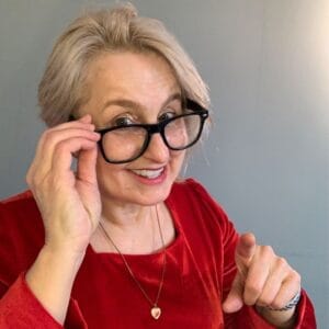 Liefdescoach Christine Filip, met lichte huidskleur met rode jurk, kijkt lachend over de glazen van de zwarte mock-up bril heen naar de camera en wijst met de linkerwijsvinger naar de camera Ze heeft een ketting met een hartje als hangertje.