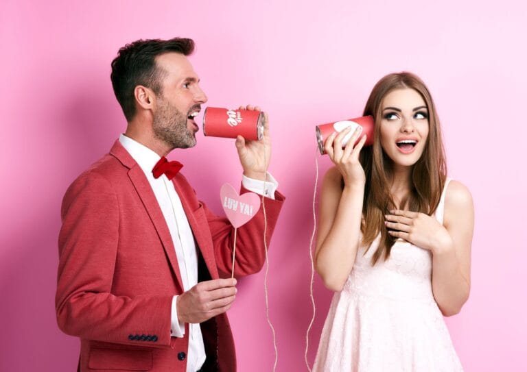 Roze achtergrond. Man en vrouw met lichte huidskleur. Man praat in een blik met een touwtje dat verbonden is met een blik dat de vrouw tegen haar oor houdt Man heeft een rood pak aan en de vrouw een lichtroze jurk.